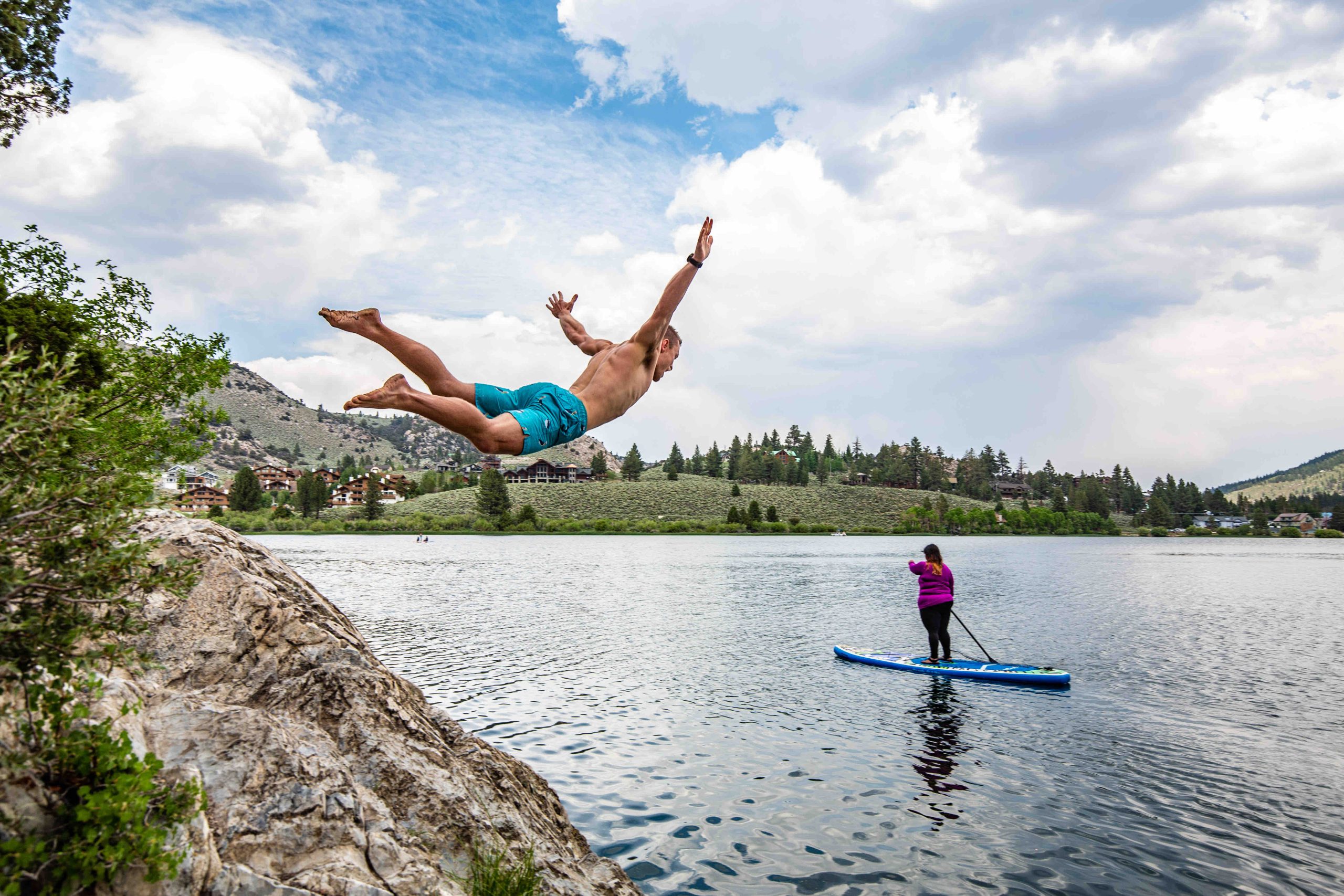 A cliff jumper mid-air over June Lake with a paddleboarder watching from below